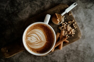 Cup of coffee with latte art on a wooden board, accompanied by cinnamon sticks, nuts, and caramel