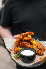 A person holding a plate of crispy, sauced chicken wings garnished with herbs. A small bowl of creamy dip accompanies the dish, highlighting a delicious and appetizing presentation.