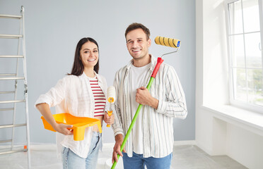 Portrait of smiling happy couple looking at camera holding paint rollers going to paint the wall of...