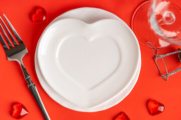Top view of white festive heart shaped ceramic empty plate served on bright red background with tableware of fork, glass and holiday decoration used for celebration of valentines day or romantic lunch