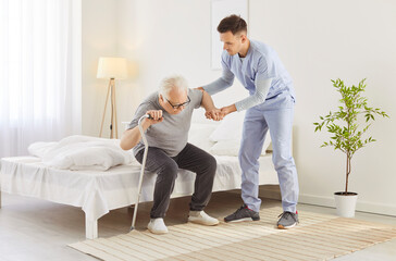 Fototapeta premium Caregiver helping an elderly patient. Young carer in a uniform helps an old man who has rheumatism, arthritis, or osteoporosis to get up from bed in the morning. Senior assistance and support concept