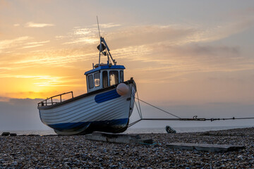 Sunrise over a single beach: Fishing boats rest on the shingle beach, bathed in the warm glow of the morning light, as the serene coastal landscape awakens