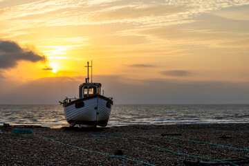 Sunrise over a single beach: Fishing boats rest on the shingle beach, bathed in the warm glow of the morning light, as the serene coastal landscape awakens