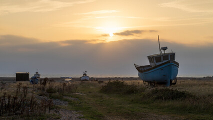 Sunrise over a single beach: Fishing boats rest on the shingle beach, bathed in the warm glow of the morning light, as the serene coastal landscape awakens