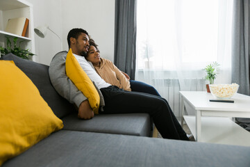 Happy African American couple enjoys quality time together while watching television. Man and woman seated closely on the couch, showcasing comfort and connection in a cozy home setting. TV, series
