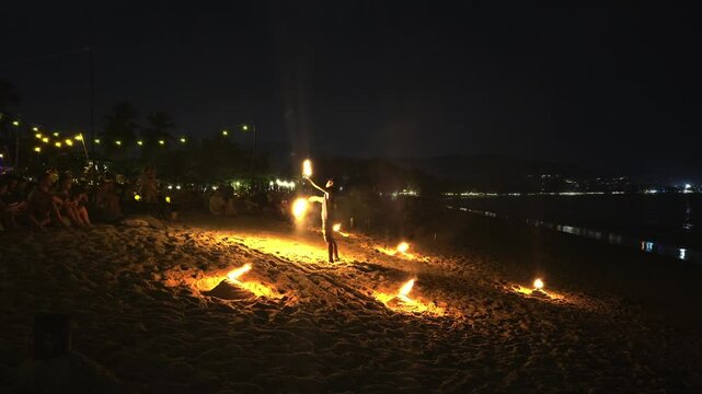 Performers manipulate flaming objects, creating light patterns on a beach at night. Spectators and string lights enhance the festive atmosphere.