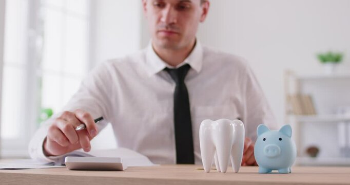 Focused man in shirt sitting at desk with small piggy bank and ceramic tooth model, using calculator, counting money, planning budget and placing coins into money bank. Savings for dental treatment.