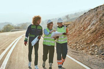 Obraz premium Three engineers, including a Muslim woman, walk together in the early morning, reviewing and analyzing wind turbine project plans while discussing innovative solutions on site.