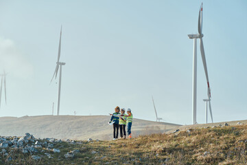 A group of engineers and environmental experts stand on a wind farm site, analyzing plans and discussing sustainable energy solutions, surrounded by wind turbines in a rural landscape