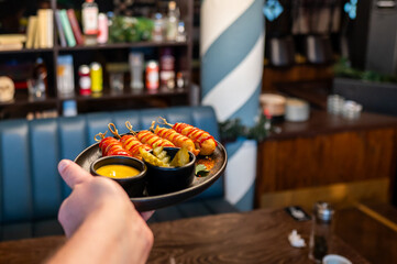 A hand holding a black plate with skewered snacks drizzled with sauce, accompanied by a small bowl of dipping sauce and seasoned vegetables, set in a cozy restaurant atmosphere.