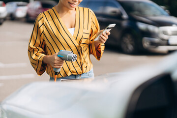 Close up of African American woman charging electro car at the electric station, using smartphone