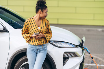 African American woman using smartphone while charging the vehicle outdoor