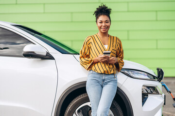 Happy African American woman holding smartphone, charging electric car, looking at camera