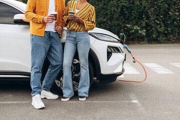 Young couple recharging electric vehicle from EV charging station, holding smartphone and coffee