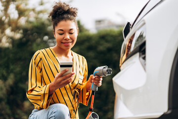 Happy African American woman using smartphone and charging car at vehicle charging station