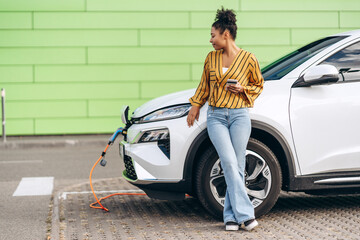 African American woman charging her electric car and using smartphone in the city