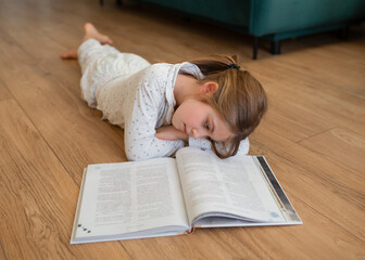 A cheerful girl in pajamas lying on the floor and enthusiastically reading a book at home. Cozy...