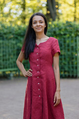 young African American woman in burgundy maxi dress is adorable female model posing in park or country house. happy lady smiles twirls in beautiful outfit on green hedge. full growth, Looks at camera