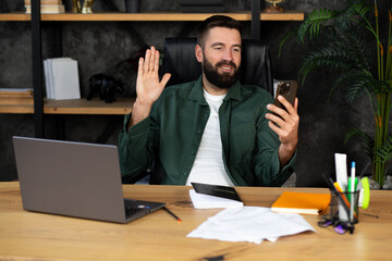 Smiling young man talking on a video call on phone, sitting in the office and gesturing with his...