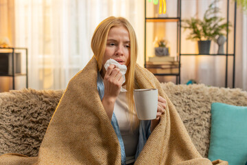 Young woman hides under blanket at home, sipping warm tea and wiping nose with tissue. Caucasian...