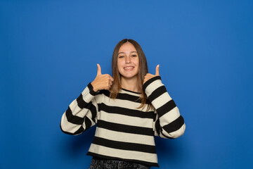 Hispanic teenage girl about 15 years old wearing striped sweater standing over isolated blue wall...