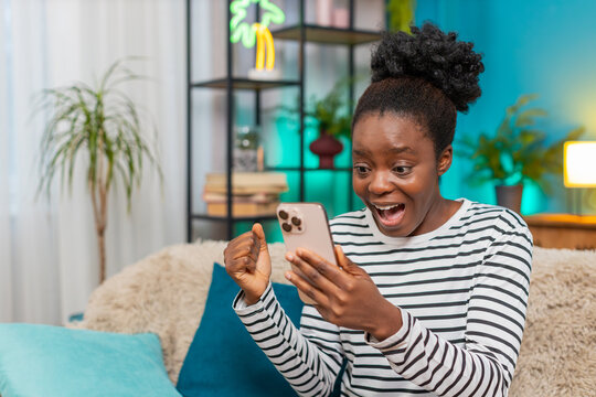 African American woman celebrating good news on smartphone. Sitting at home sofa, joyful expression, happy emotions, receiving message or winning in lottery, engaging indoor atmosphere, excited mood