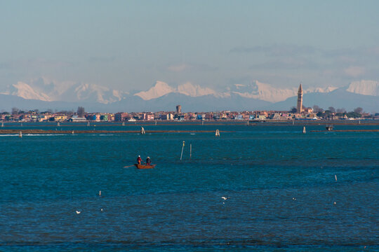 Panorama dell&rsquo;isola di Burano con le Dolomiti innevate sullo sfondo e una barca a remi rossa che naviga nella laguna
