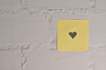 Yellow sticker paper with small black heart mark in the side on white brick wall background. Romantic message for Valentine's Day