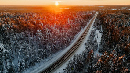 The car is driving along the road among snow-covered fir trees in a winter forest at sunset