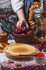 Pancakes on the table with traditional patterns, next to the samovar and caviar. From behind, a woman's hand takes an apple from a basket