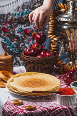 Pancakes on the table with traditional patterns, next to the samovar and caviar. From behind, a woman's hand takes an apple from a basket