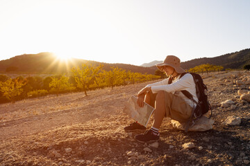 Female hiker sitting on rocky terrain, examining map during golden-hour sunlight with mountainous...