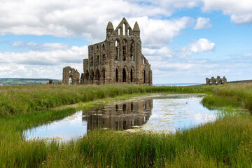 Obraz premium Former Christian monastery now Benedictine Whitby Abbey Church in Whitby, UK on the East Cliff along the coast with reflection of the building in the pond against a white clouded blue sky