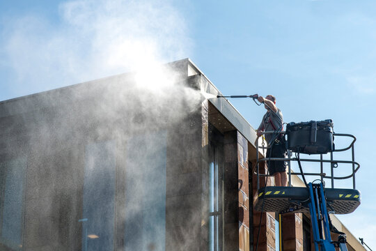 Man standing on aerial work platform or cherry picker with high pressure water gun cleaning the fa&ccedil;ade of a modern building with water spray against sun covering part of image