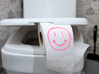 Close-up a roll of toilet paper with a funny smiley face drawn on it lies on the toilet lid in the...
