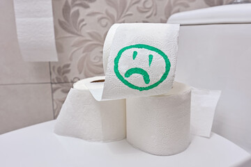 Close-up a several rolls of toilet paper with a sad smiley face drawn green color on it lies on the toilet lid in the apartment's bathroom.
