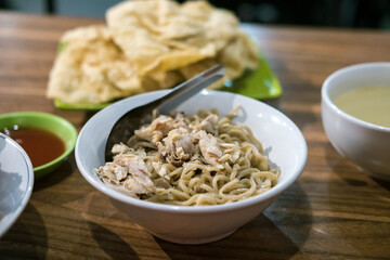 A bowl of Mie Ayam (chicken noodles) with chopped chicken, a spoon resting on top, accompanied by crackers, broth, and chili sauce.