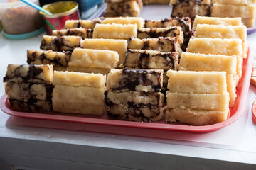 Sliced kue pukis (Indonesian mini pancakes) with chocolate and plain flavors, neatly arranged on a pink tray at a street food stall. Kue pukis is a traditional Indonesian snack made from flour, eggs, 