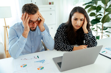 Stressed colleagues analyzing data and charts on a laptop and printed sheets at a desk, working...