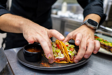 A chef carefully garnishes two delicious tacos with fresh ingredients, served on a black plate alongside a small bowl of sauce. A vibrant kitchen background adds to the culinary atmosphere.