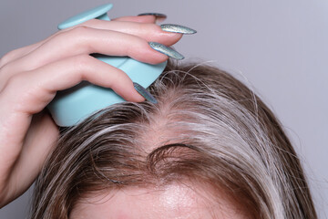 Naklejka premium Chestnut hair with gray strands is gently massaged by a woman's hand holding a blue massager. The massage with serum soothes the scalp and strengthens hair follicles.