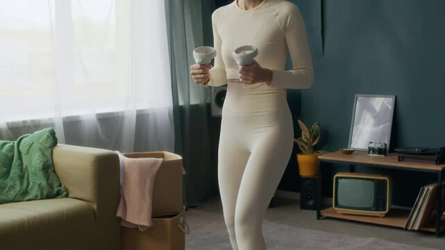 Tilt up slow motion shot of cheerful gen Z girl in white activewear playing virtual reality game in wireless goggles in living room in retro style