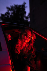A Stylish Woman in Neon Light by a Car at Dusk, Showcasing Her Unique Style and Charisma