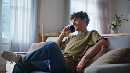 Fototapeta premium Smiling man talking cellphone at living room couch closeup. Guy communicating