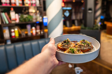 A hand holding a plate of gourmet food featuring topped toast rounds, placed in a stylish restaurant setting with a blurred background of shelves and decor. Perfect for food or dining themes.