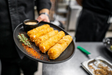 A plate of crispy golden-fried cheese sticks served with a small bowl of dipping sauce, garnished with fresh thyme and a sprinkle of red spice, in a professional kitchen setting.