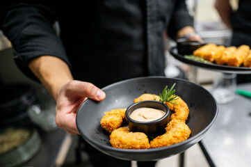 A close-up of a chef serving a black plate with crispy fried shrimp bites and a small cup of dipping sauce. The background is softly blurred, focusing on the delicious dish.