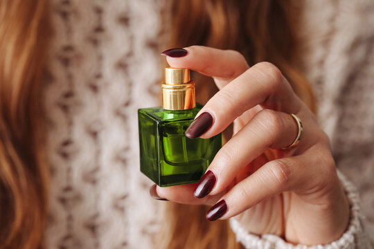 Woman in sweater holding green perfume bottle. Fragrance spray lifestyle background. Female scent application. Using perfume by a girl. Fragrance in glass bottle in fingers.