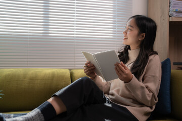 Young woman wearing casual clothes sitting on a comfortable sofa in her living room taking notes in...