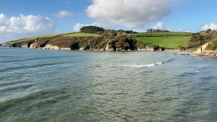 Fototapeta premium A tranquil coastal scene at Mothecombe Beach, Devon, showcasing calm waves, surfers, green cliffs, and a clear, sunny sky. Perfect for concepts related to nature, travel and the seaside - UK